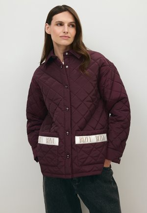 Vrouw draagt een donkerpaarse doorgestikte jas met opgestikte zakken met label "BIZUU" en een donkere broek, staand tegen een effen lichte achtergrond.