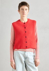 Κόκκινο πλεκτό γιλέκο με στρογγυλή λαιμόκοψη και μαύρα κουμπιά, τοποθετημένο πάνω από ένα μακρυμάνικο πουκάμισο με ρίγες. Μαλακή υφή, άνετη εφαρμογή και φωτεινό χρώμα.