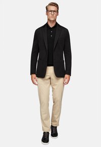 Black textured blazer over a black polo, paired with beige trousers and black sneakers. Clear glasses accentuate the contemporary look.