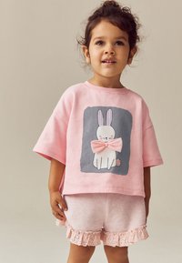 T-shirt oversize rose clair avec un motif de lapin blanc portant un nœud rose. Associé à un short rose avec une bordure en dentelle à l'ourlet.