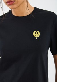 T-shirt en coton noir avec un graphique de visage souriant jaune avec des gouttes, situé sur la partie gauche de la poitrine ; coupe décontractée et manches courtes.
