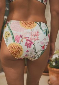 Blommiga bikiniunderdelar med vit bakgrund och färgglada blommönster, inklusive gula och rosa blommor, med en jämn tygtextur.