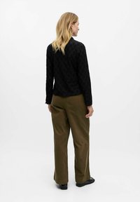Chemisier noir en dentelle à manches longues avec une texture à motif, associé à un pantalon large vert olive. Des chaussures plates noires complètent la tenue.