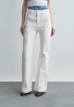 Białe, wysokociśnieniowe dzwonowe dżinsy wykonane z denimu, z przednimi kieszeniami, zapięciem na guziki oraz gładką fakturą. Szare buty z wąskim noskiem.