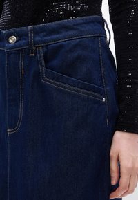 Σκούροι τζιν παντελόνια με χρυσή ραφή, λεπτομέρεια μπροστινής τσέπης και κούμπωμα με κουμπί. Το υλικό φαίνεται ανθεκτικό με λεία υφή.