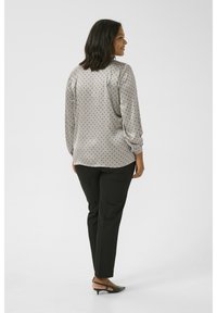 Blouse gris clair à pois noirs, à manches longues et avec des poignets élastiques, associée à un pantalon noir fuselé et des chaussures ouvertes noires à talon.