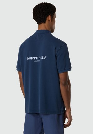 Hombre con cabello corto y rizado, vistiendo una camiseta polo azul marino con la inscripción "NORTH SAILS ARCHIVE" en la espalda, sobre un fondo liso.