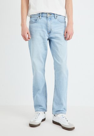 Lyseblå rettskårne jeans i falmet denim med frontlommer, glidelås, og en messingknapp-lukking. Brukt med hvite sneakers.