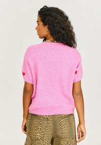 Ružový chlpatý sveter s krátkymi rukávmi, s červeným srdcom na ľavom rukáve. Skombinovaný s béžovými nohavicami s leopardím vzorom, hladká textúra.