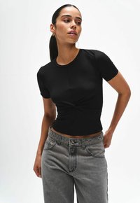 Zwart cropped t-shirt met korte mouwen, voorzien van een knoopdetail aan de voorkant. Gemaakt in combinatie met lichtgrijze high-waisted jeans, met een gladde stofstructuur.