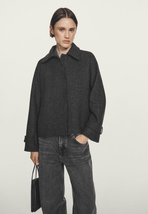 Donkergrijze wollen jas in een mix met een kraag, knoopsluiting en opgerolde manchetten, gecombineerd met hooggesneden grijze denim broek en een kleine handtas.