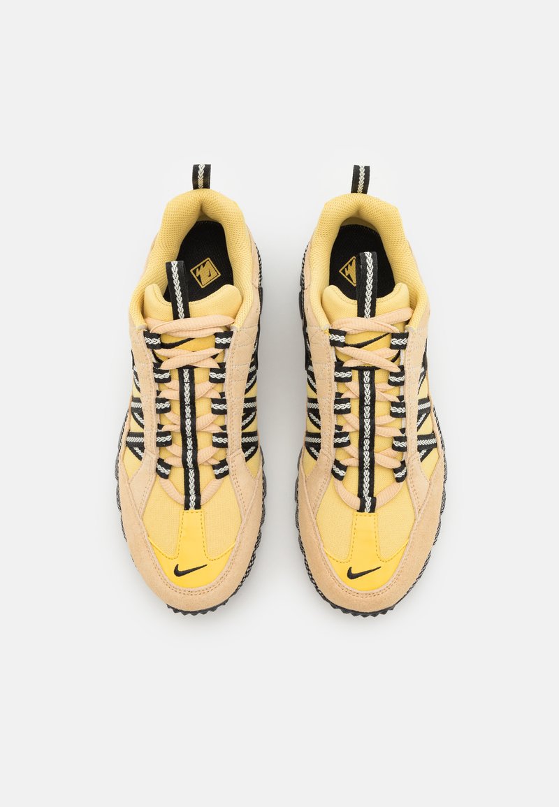 Des sneakers jaunes et beiges avec des accents noirs, présentant un dessus texturé, des lacets à motifs noir et blanc, et un logo Nike noir sur le côté.