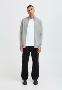Chaqueta gris con cierre, hecha de un tejido suave, combinada con una camiseta blanca y pantalones negros. El modelo se encuentra en una postura y expresión neutra.