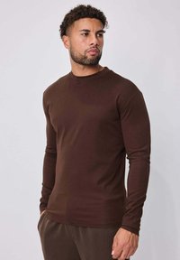 Chemise marron à manches longues en tissu lisse, avec un col rond et une coupe ajustée, accompagnée d'un pantalon marron assorti.