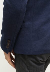 Marinblazer i texturerat tyg. Har fyra bruna knappar vid ärmslut och en skräddarsydd passform. Synlig söm vid axeln.