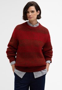 Roter strukturierter Strickpullover mit Rundhalsausschnitt und gerippten Bündchen, über einem gestreiften Hemd mit Kragen getragen, kombiniert mit dunkelblauen Hosen.
