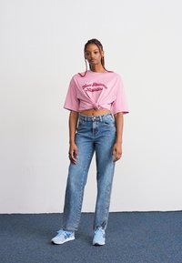 Young woman stands against a plain wall wearing a knotted pink New Balance top, high-waisted blue jeans, and light blue sneakers.