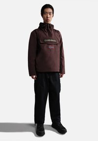 Giacca a pullover marrone con cappuccio, tasca frontale con logo e patch della bandiera. Abbinata a pantaloni cargo neri e scarpe sportive nere.