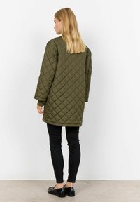 Gequiltede olijfgroene jas met lange mouwen en een relaxte pasvorm, gecombineerd met zwarte skinny jeans en glanzende zwarte schoenen.