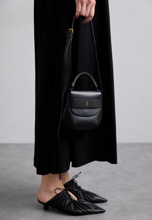 Pequena mala de couro preto com forma arredondada, pega em cima e alça removível. Usada com vestido de tecido preto e sapatos pretos plissados.