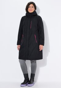 Musta vedenpitävä takki hupulla, jossa on vaaleanpunaisia vetoketjuaksentteja, pitkät hihat ja suora leikkaus. Vaatteen päällä harmaat farkut ja mustat korkeat saappaat.