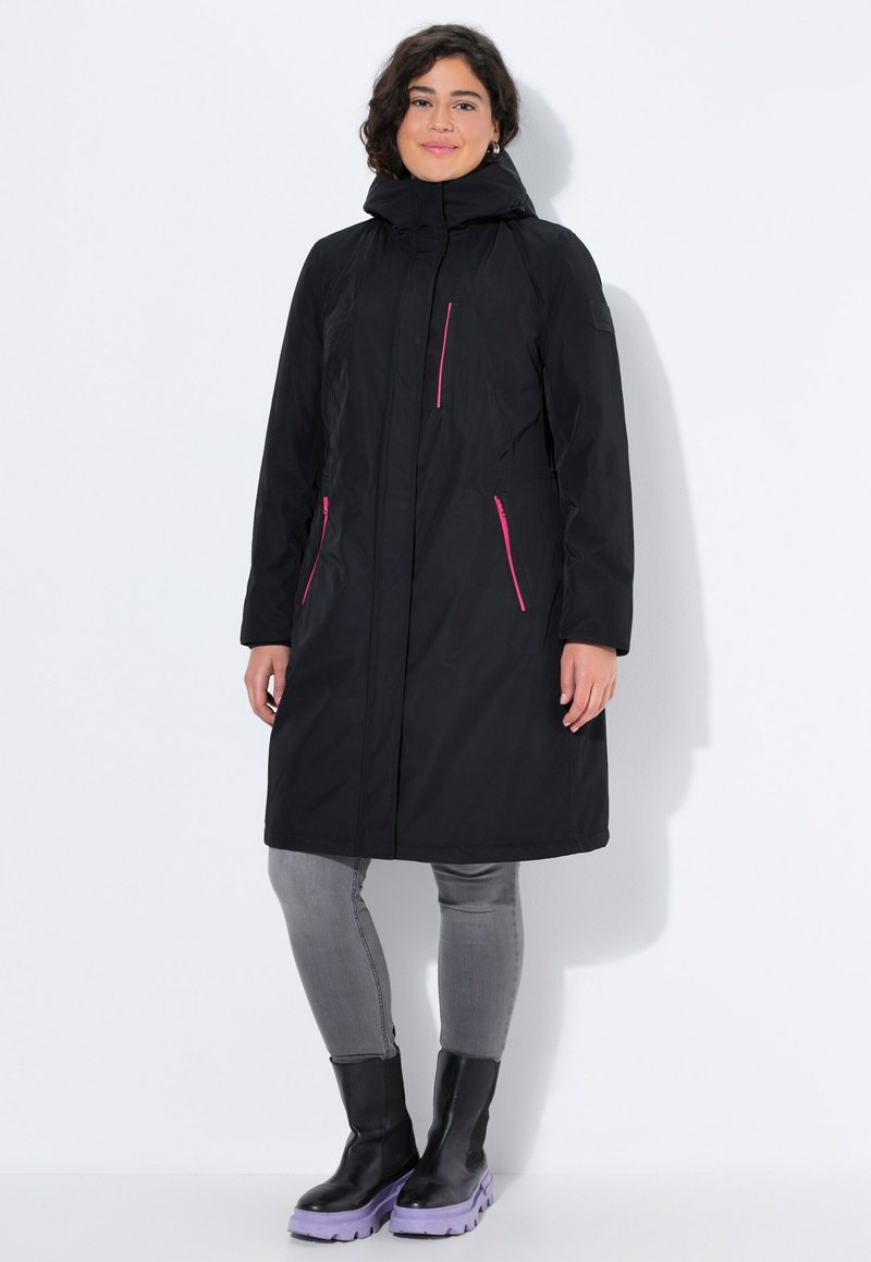 Musta vedenpitävä takki hupulla, jossa on vaaleanpunaisia vetoketjuaksentteja, pitkät hihat ja suora leikkaus. Vaatteen päällä harmaat farkut ja mustat korkeat saappaat.