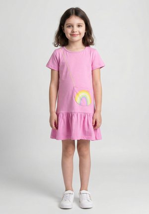Młoda dziewczynka w różowej sukience z kieszenią ozdobioną tęczowym naszywką oraz białych trampkach z tęczowymi detalami, stojąca na jednolitym tle.