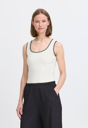 Ärmelloses cremefarbenes Tanktop mit weitem Ausschnitt und schwarzem dreieckigem Besatz. Kombiniert mit strukturierten schwarzen weit geschnittenen Hosen.