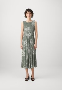 Robe midi florale verte avec des fleurs blanches, sans manches, jupe plissée et taille cintrée. Portée avec des talons ouverts noirs.