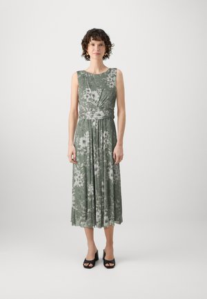 Abito midi floreale verde con fiori bianchi, senza maniche, gonna plissettata e vita aderente. Indossato con tacchi neri open-toe.