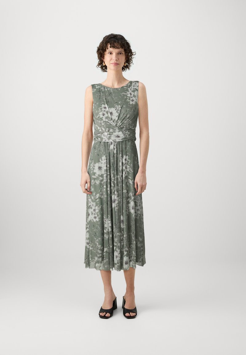 Robe midi florale verte avec des fleurs blanches, sans manches, jupe plissée et taille cintrée. Portée avec des talons ouverts noirs.