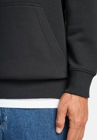 La mano di un uomo riposa di lato, indossando una felpa nera con tasca frontale, una maglietta bianca e jeans blu.