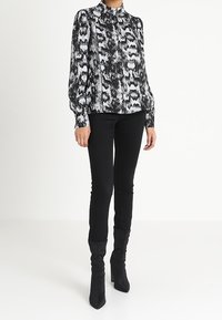 Blouse à imprimé serpent noir et blanc avec col montant, manches bouffantes et tissu léger, associée à un pantalon noir ajusté et des bottes à talons.