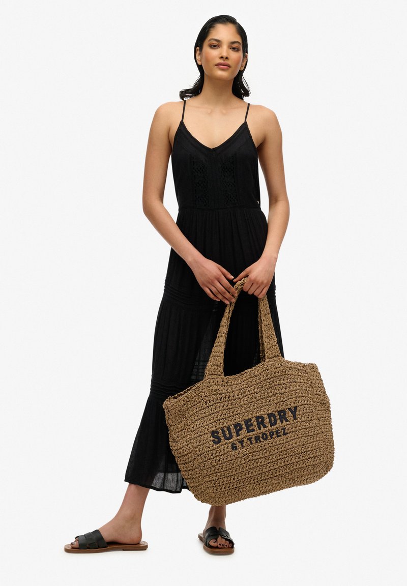 Bolso tote trenzado de paja con la inscripción "Superdry St. Tropez" en negro, complementado con un vestido negro fluido con tirantes ajustables y diseño de capas.