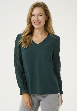Top teal con scollatura a V e maniche lunghe, caratterizzato da dettagli in pizzo intricati. Il tessuto è morbido e ha una vestibilità rilassata. Abbinato a jeans grigi chiari.