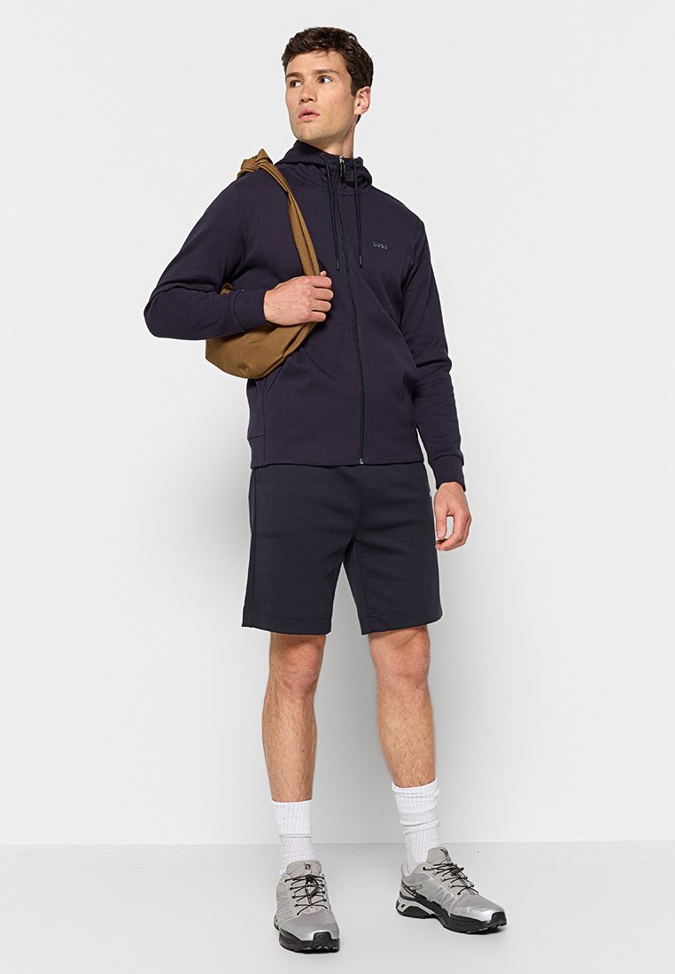 Felpa zip-up blu scuro con collo alto, abbinata a pantaloni corti grigi. Completata da una borsa a tracolla marrone chiaro e scarpe sportive grigie.