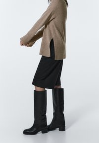 Osoba w beżowym swetrze, czarnej spódnicy do kolan i czarnych kozakach do kolan, stoi na tle jasnego, jednolitego tła.