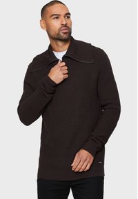 Dunkelbrauner Strickpullover mit einem breiten Kragen und einem Reißverschluss; strukturiertes Material, figurbetonte Ärmel und ein Logo-Detail am Saum.