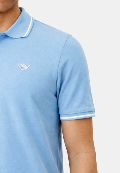 Homme portant un polo bleu clair à manches courtes avec bordure blanche sur le col et les extrémités des manches, et petit logo blanc brodé sur la poitrine.