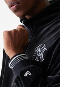 Veste en velours noir avec un col haut, ornée d'un logo brodé blanc et de rayures blanches sur les poignets. Gros plan d'un poing serré.