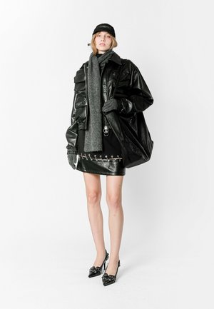 Chaqueta de cuero negra de gran tamaño sobre una falda corta negra con detalles plateados, combinada con una bufanda y guantes grises, finalizando con tacones negros.