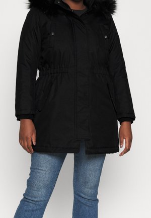 Personne portant un manteau d'hiver noir avec une capuche bordée de fourrure et un jean bleu, debout devant un fond clair uni.