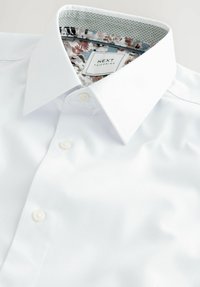 Camisa blanca de botón con tejido texturizado, que presenta un interior del cuello con un patrón floral y una etiqueta con el diseño "NEXT TAILORING".