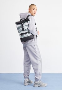 Zaino grigio con motivo camouflage, dettagli neri, chiusura roll-top e spallacci regolabili, abbinato a una felpa grigia e pantaloni coordinati.