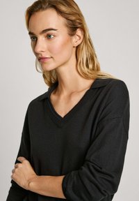 Melns adīts džemperis ar v-veida kaklu un īsām, sarullētām piedurknēm. Gluda tekstūra, ar smalku apkakles dizainu.
