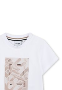 T-shirt bianco a maniche corte con un motivo geometrico beige stampato sul davanti. Presenta bottoni a pressione sulla spalla per facilitare la vestizione.