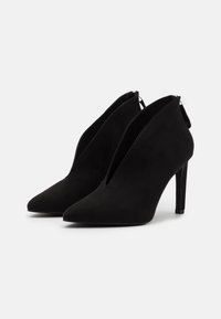 Botas de ante negras hasta el tobillo con punteras puntiagudas, cierres de cremallera en la parte posterior y tacones de aguja de 10 cm. Textura lisa mate, diseño minimalista.