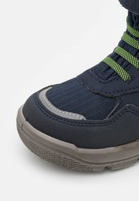 Blauwe textielschoen met een gestructureerde donkergrijze rubberen zool, groene veters en versterkte neus. Close-up van het ontwerp dat de details benadrukt.