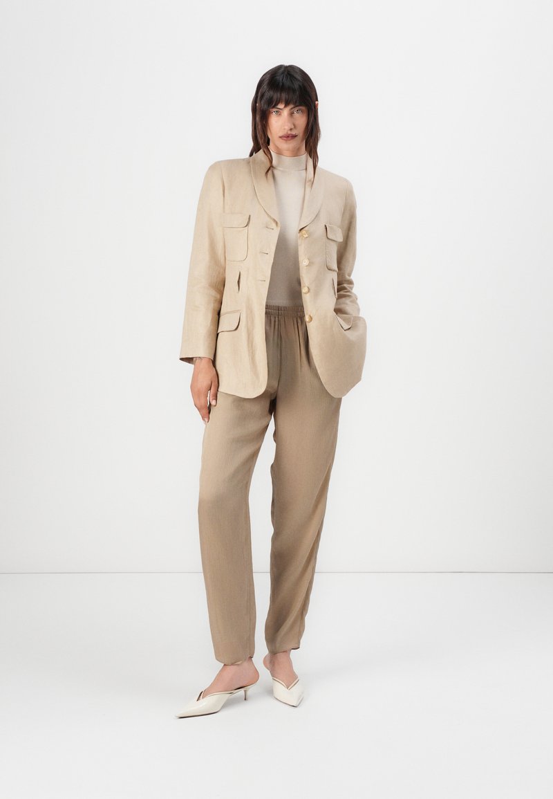 Beiger Leinen-Blazer mit vier Fronttaschen, goldenen Knöpfen und passenden beigen Plisseehosen. Weiße spitze Mules vervollständigen das Outfit.