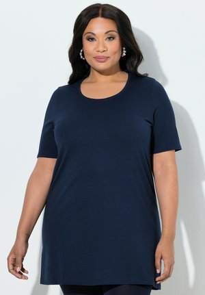 Kvinde med mørkt, bølget hår, iført en navyblå tunikaskjorte med korte ærmer og perle-huløreringe, stående foran en lys baggrund.